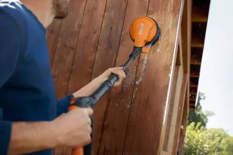 Hombre usando un accesorio de cepillo para limpiar la fachada de madera de su casa.