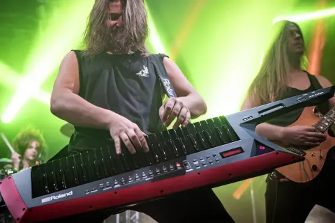 Muzyk gra na keyboardzie na pasku, podczas gdy gitarzysta i perkusista tworzą rockową atmosferę.