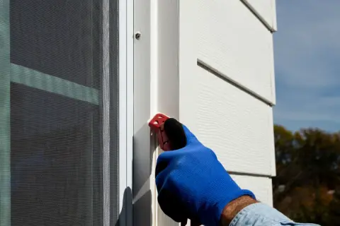 A gloved hand uses a red tool to smooth caulk along a window frame, demonstrating how to caulk windows for a weather-tight seal.