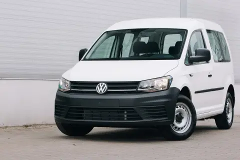 Biały Volkswagen Caddy, samochód osobowy zarejestrowany jako ciężarowy bez kratki, stoi przed szarą ścianą.