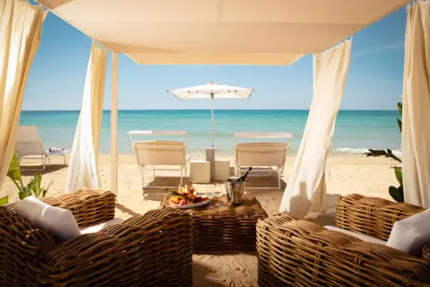 Lussuosa area lounge con divani in vimini, tavolino con frutta e champagne, lettini e ombrellone su una spiaggia di sabbia dorata. Un'oasi tra i migliori beach clubs Puglia.
