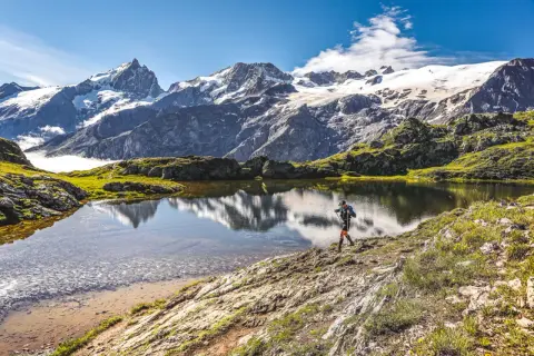 Un randonneur traverse le plateau d'Emparis, un lac alpin reflétant des montagnes enneigées sous un ciel bleu.