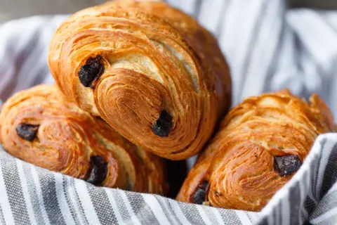Tre deliziosi pain au chocolat dorati, con strati sfogliati e gocce di cioccolato fondente, adagiati su un canovaccio a righe.