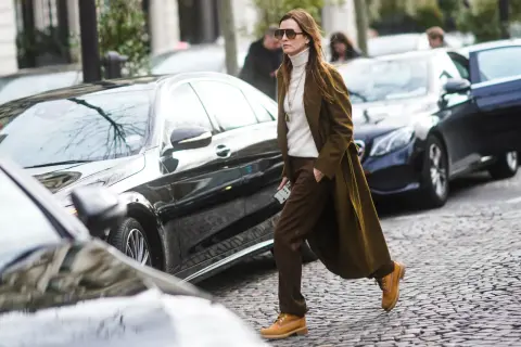 Donna con un elegante outfit con Timberland, cappotto verde oliva, dolcevita bianco e pantaloni marroni, cammina su un acciottolato.