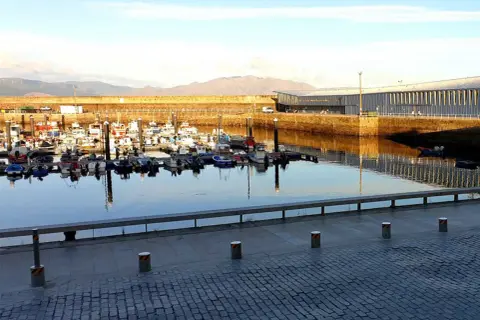 Barcos pesqueros amarrados en el puerto de Finisterre, con un edificio moderno al fondo y montañas en la distancia.