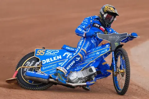 Bartosz Zmarzlik w niebieskim kevlarze z numerem 95, na motocyklu Orlen, ściga się na żużlu. Jego wzrost nie przeszkadza w dynamicznej jeździe.