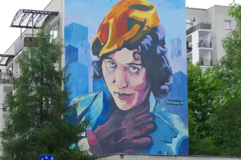 Mural przedstawiający Zofię Stryjeńską w żółtym beretce, z dłonią na piersi. Tło to błękitne, geometryczne budynki.