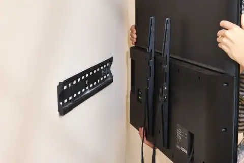 Hands carefully lift a TV, showing how to remove TV from wall mount. The wall bracket is visible.
