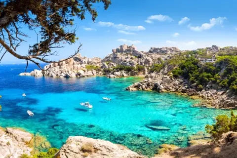 Turkusowa woda zatoki na Costa Smeralda, łodzie kołyszące się na falach, skaliste wybrzeże porośnięte zielenią.