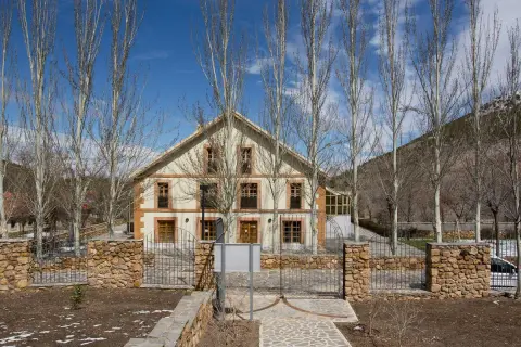 Edificio de piedra y madera con árboles secos delante. El camino de entrada está pavimentado con piedras. Las menas de la montaña se ven al fondo.