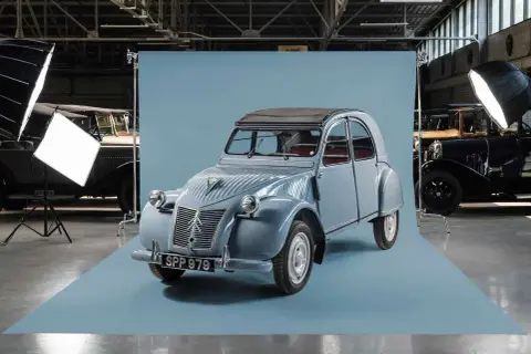 En ljusblå Citroën 2CV står i fokus, redo för fotografering. Bakom den syns andra klassiska bilar och studiobelysning.