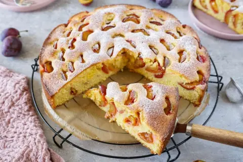 Saftiger pflaumenkuchen mit gefrorenen pflaumen, bestäubt mit Puderzucker. Ein Stück wird gerade herausgenommen.