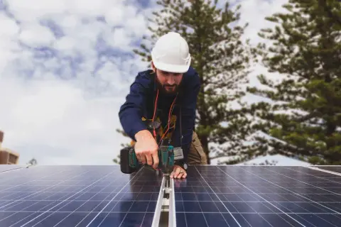 Técnico instala paneles solares, cumpliendo con el real decreto autoconsumo. Edificio y árboles al fondo.