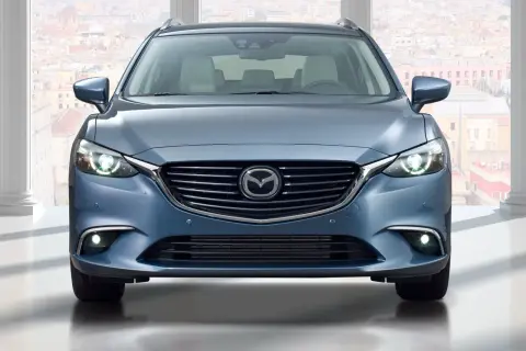 Niebieska Mazda 6 GJ kombi z przodu, z włączonymi światłami, na tle miasta.