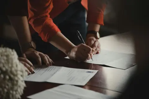 Dłonie podpisują dokumenty na drewnianym stole. Jedna z osób ma na nadgarstku zegarek, a druga nosi bransoletkę. W tle widać fragment pomponu. Klauzula RODO jest ważnym elementem umowy.