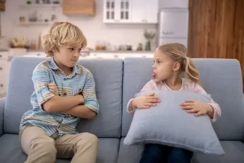 Bambini che litigano sul divano. Lui incrocia le braccia, lei fa la linguaccia con un cuscino.