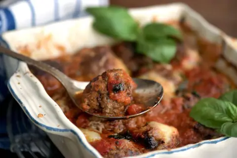 En sked lyfter en köttbulle täckt av mustig tomatsås ur en ugnsform. En härlig pasta med tomatsås väntar!