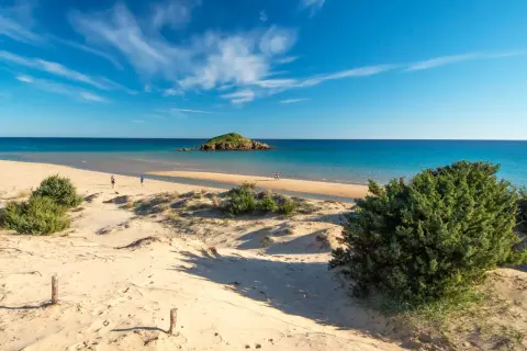 Dune sabbiose e mare turchese, un'isola rocciosa in lontananza. Un assaggio delle spiagge più belle Sardegna, con persone che passeggiano sulla riva.