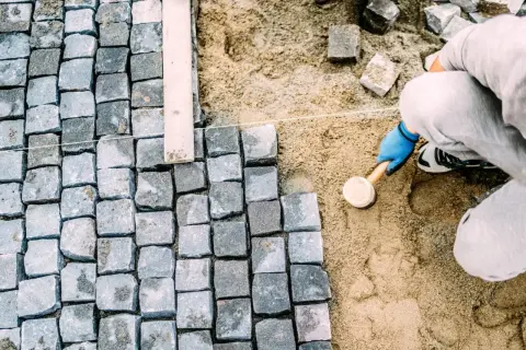 Pracownik w niebieskich rękawiczkach ubija kostkę brukową gumowym młotkiem, tworząc równą nawierzchnię.