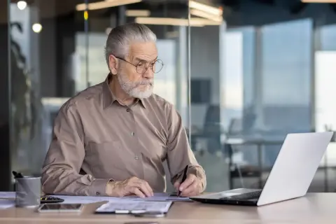Starszy mężczyzna w okularach, z siwą brodą, siedzi przy biurku i pracuje. To jego praca na emeryturze, gdzie nadal jest aktywny zawodowo.