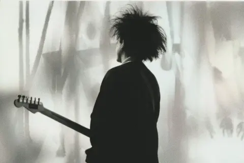 Chłopcy nie płaczą. Robert Smith z The Cure z gitarą, w cieniu, z charakterystyczną fryzurą.