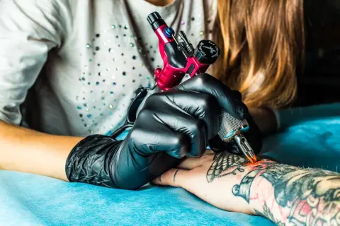 Künstler mit schwarzen Handschuhen arbeitet an einem Tattoo auf dem Arm. Ein rotes Tattoo-Gerät wird verwendet, um ein neues Design zu erstellen.