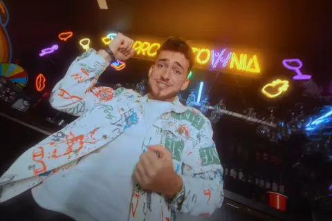 Hej, co zrobiłaś? Jaka głupia byłaś! Młody mężczyzna w kolorowej kurtce tańczy na tle neonów i baru.