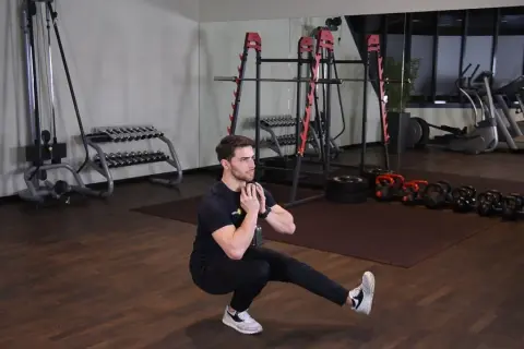 Mężczyzna wykonuje ćwiczenie pistol squat z hantlem. W tle siłownia z maszynami i ciężarami.