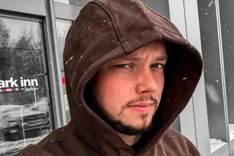 Mężczyzna w brązowej bluzie z kapturem, na tle drzwi hotelu Park Inn. Widać padający śnieg.