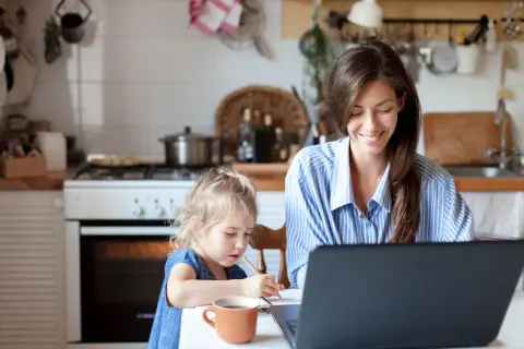 Frau arbeitet am Laptop, Kind malt daneben. Ein Beispiel für Home Office Sicherheit, wo Familie und Beruf harmonisch vereint werden.