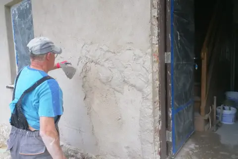 Pracownik tynkuje stare ściany zewnętrzne, nakładając zaprawę kielnią. Widać fragment drzwi i schody w tle.