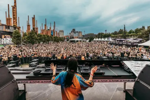 DJ set al futur festival, folla in delirio sotto un cielo nuvoloso, con un'architettura industriale sullo sfondo.