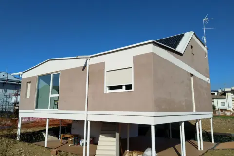 Casa moderna con intonaco argilla, pannelli solari e ampie vetrate. La struttura è rialzata su pilastri bianchi.