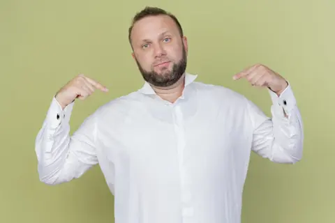 Mann zeigt auf sich und sein weißes Hemd. Er fragt sich, welches Unterhemd unter dem Hemd am besten passt.