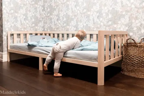 A toddler climbs into a Montessori bed, showcasing its low profile and ideal dimensions for independent exploration.