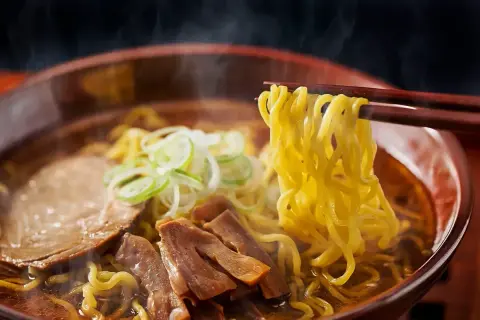 En skål med ångande ramen, med en skillnad på nudlar och pasta som tydligt syns. Nudlarna lyfts med ätpinnar, med kött och grönsaker i buljongen.