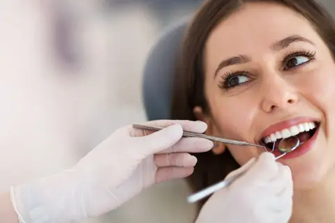 Uśmiechnięta pacjentka podczas zabiegu sanacji jamy ustnej. Dentysta bada zęby lusterkiem.
