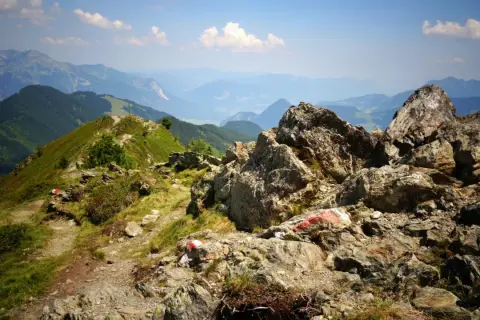 Senderos marcados con rojo y blanco serpentean por las laderas verdes de los Picos de Europa en verano, con rocas imponentes y un valle brumoso al fondo.