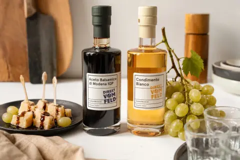 Zwei Flaschen Balsamico und Condimento Bianco, daneben Käsewürfel mit Balsamico und Trauben.