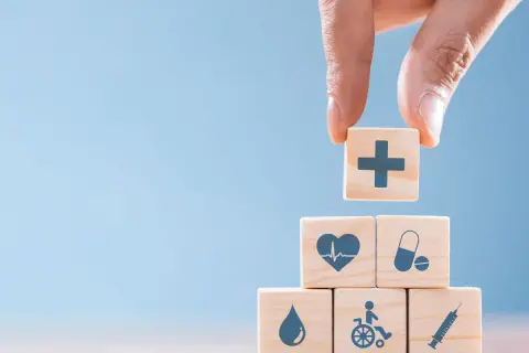 Dłoń układa kostkę z symbolem krzyża na piramidzie z kostek z ikonami zdrowia i bezpieczeństwa. To symboliczne przedstawienie obowiązków pracodawcy w zakresie BHP.