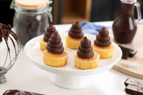 Fem cupcakes med fluffig chokladganache på en vit tårtfat. Ingredienser för chokladganache syns i bakgrunden.