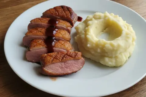 Soczysta pierś z kaczki, pokrojona w plastry, polana sosem, obok porcja purée ziemniaczanego.