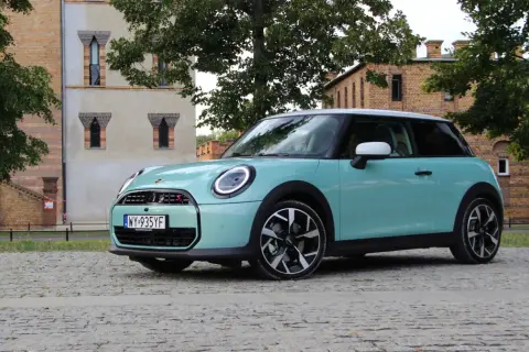 Turkusowy Mini Cooper stoi na brukowanej drodze przed ceglanym budynkiem.