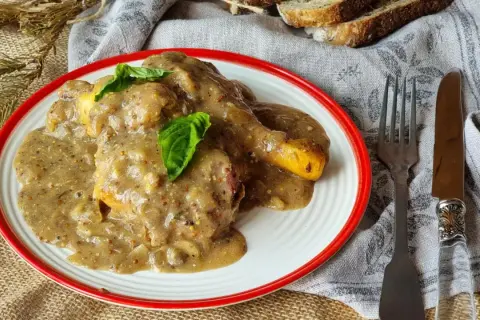Delicioso pollo a la mostaza cubierto con una cremosa salsa, servido en un plato blanco con borde rojo. Acompañado de pan y cubiertos.