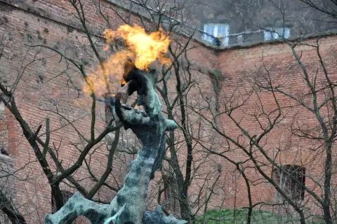 Pomnik smoka wawelskiego zieje ogniem na tle ceglanej ściany.