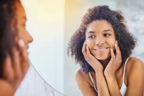 Une femme aux cheveux bouclés se regarde dans un miroir, les mains posées sur ses joues, comme pour se masser le visage. Elle sourit doucement.