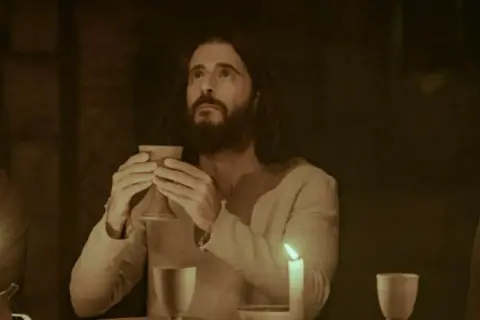 Jesus, dargestellt von Dallas Jenkins, hält einen Kelch bei einem Abendmahl. Kerzenlicht beleuchtet die Szene.