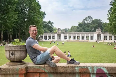 Ein Mann sitzt auf einer Mauer im Park am Stadtbad Charlottenburg Wilmersdorf. Menschen entspannen sich auf der Wiese vor dem Gebäude.