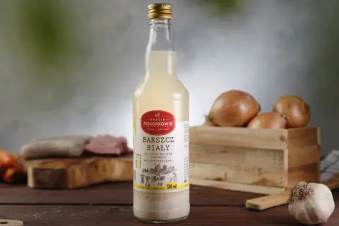 Szklana butelka z etykietą "Barszcz biały" na oryginalnym zakwasie, obok cebule i czosnek.