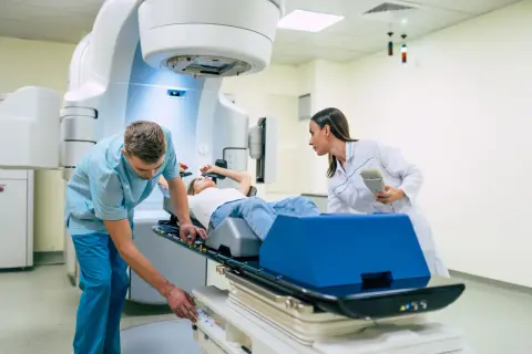 Ein Patient liegt auf einer Liege, während ein Techniker und eine Ärztin eine perkutane Strahlentherapie vorbereiten.